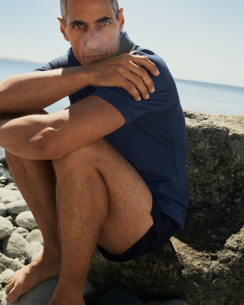 Axel Hermann in a navy polo crouches on a large rock along the coastline
