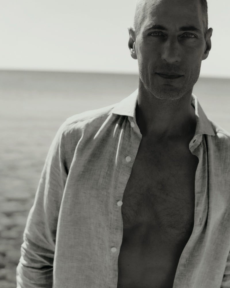 Black and white close-up portrait of Axel Hermann in an unbuttoned linen shirt with the sea behind him