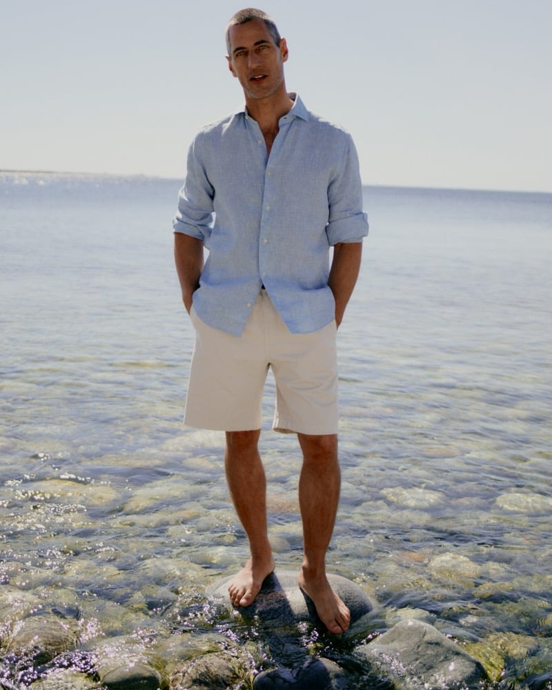 Axel Hermann in a light blue linen shirt and cream shorts standing in shallow coastal water
