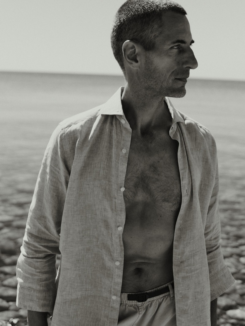 Black and white profile portrait of Axel Hermann in an open linen shirt with belted shorts, ocean in the background