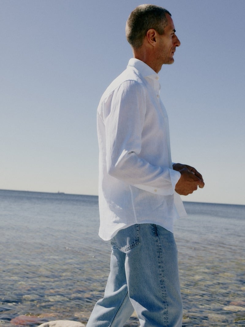 Axel Hermann in a white linen shirt and light-wash jeans walking along the shoreline