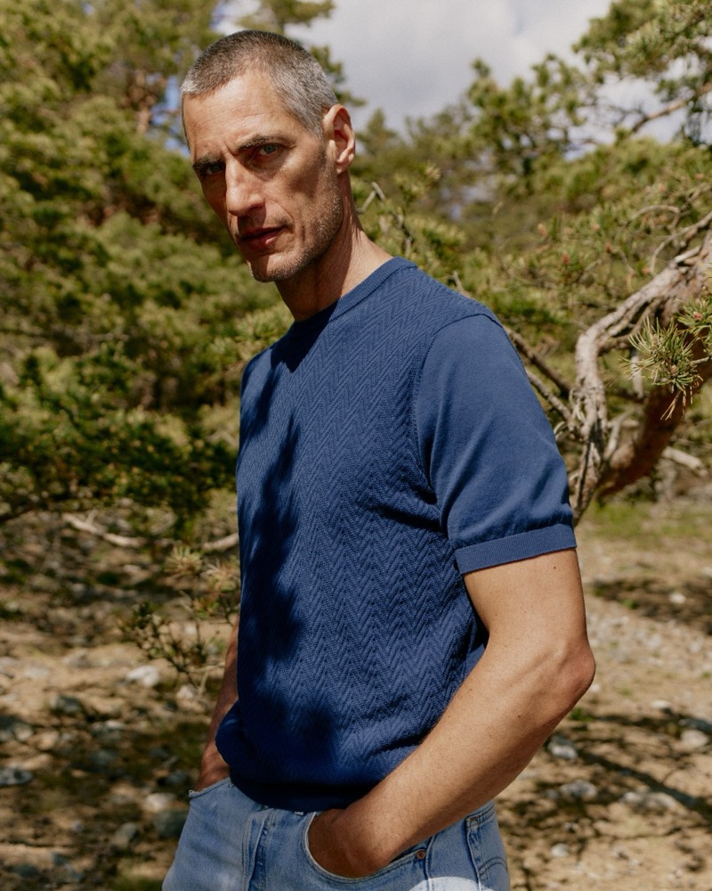 Axel Hermann in a navy herringbone-knit polo and light-wash jeans stands among pine trees