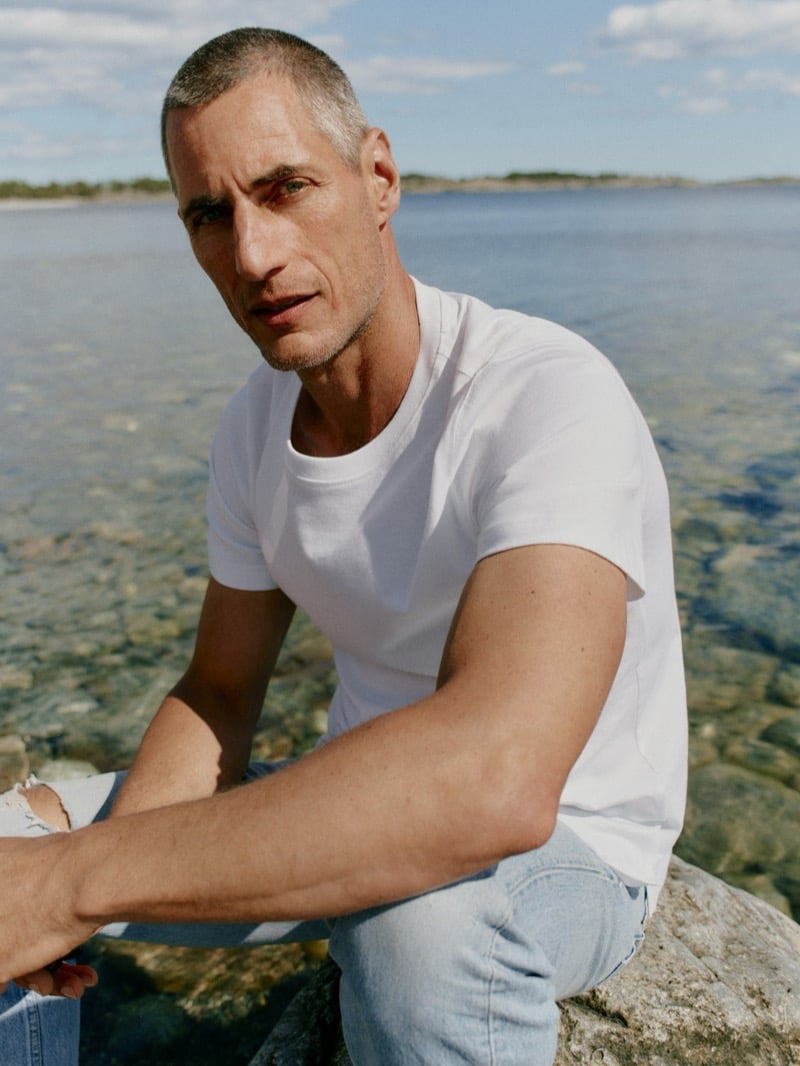 Axel Hermann in a white T-shirt seated on a rock along the water's edge