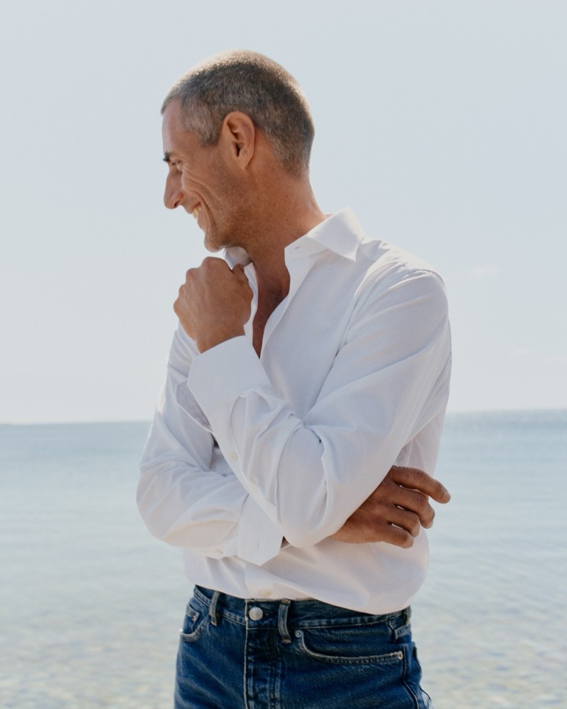 Axel Hermann in a white shirt and dark-wash jeans smiling along the shoreline