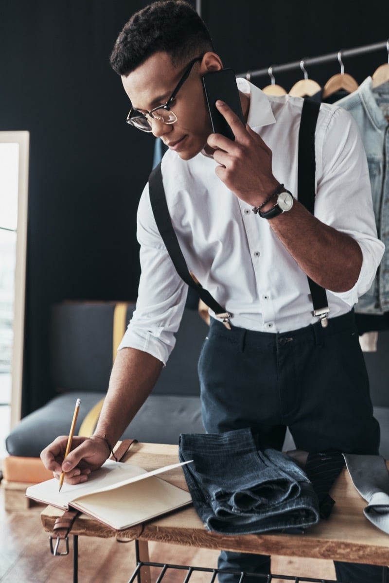 A designer takes a call while jotting notes in a sketchbook beside folded denim and ties