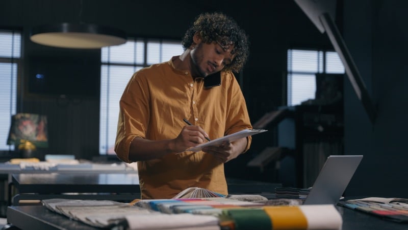 A designer takes a phone call while writing notes on a clipboard among fabric swatches