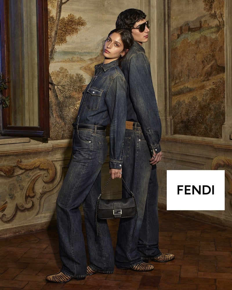 Christian Mammana in a dark-wash denim shirt and jeans stands behind a fellow model in a matching denim look, her hand resting on a Fendi Baguette bag, frescoed walls behind them