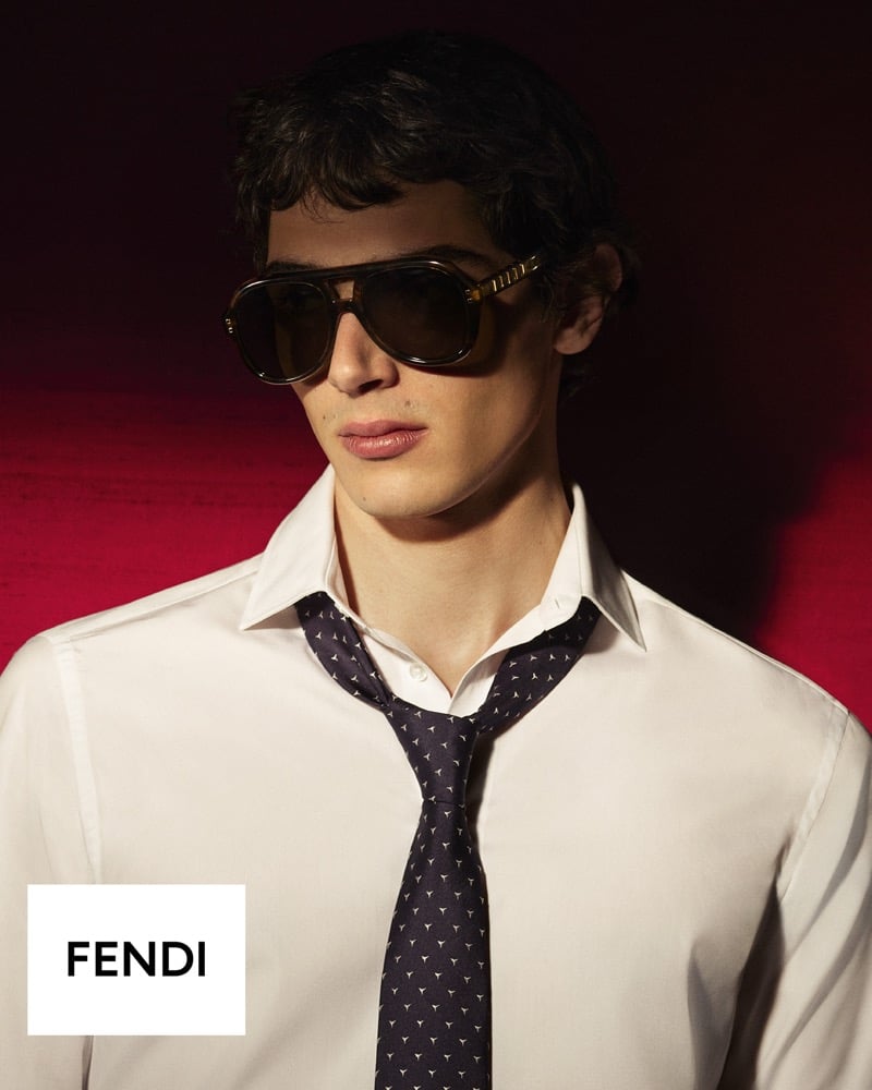 Close portrait of Christian Mammana in a white dress shirt, a dark printed tie, and tortoiseshell aviator sunglasses against a deep red backdrop