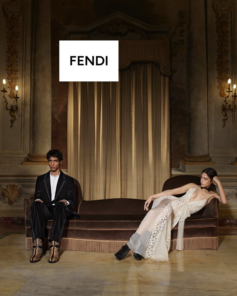 Mansur Lefqir sits upright in a black velvet tuxedo and zebra-print boots while Serena Sefyan reclines in a sheer lace gown on a neighboring velvet chaise, a Fendi logo card above them