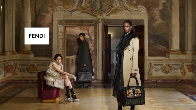 Chu Wong sits on a red chair in a metallic-embellished dress, Mansur Lefqir stands in a dark coat in the doorway behind her, and Faith Johnson stands at right in a belted trench coat with a fur collar, inside a frescoed hall