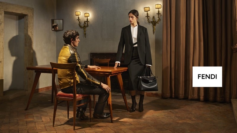 Mansur Lefqir sits at a wooden table in a black jacket with gold embroidered trim while Serena Sefyan stands beside him in a dark tailored suit, holding a Fendi handbag