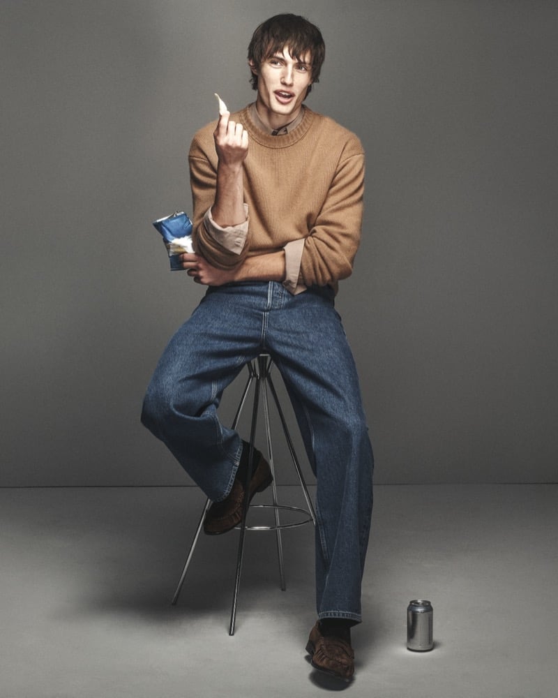 Thatcher Thornton sits on a stool in a camel cashmere crew neck sweater and blue denim jeans with brown suede loafers, snacking on a chip against a gray studio backdrop for FRAME's fall 2026 campaign