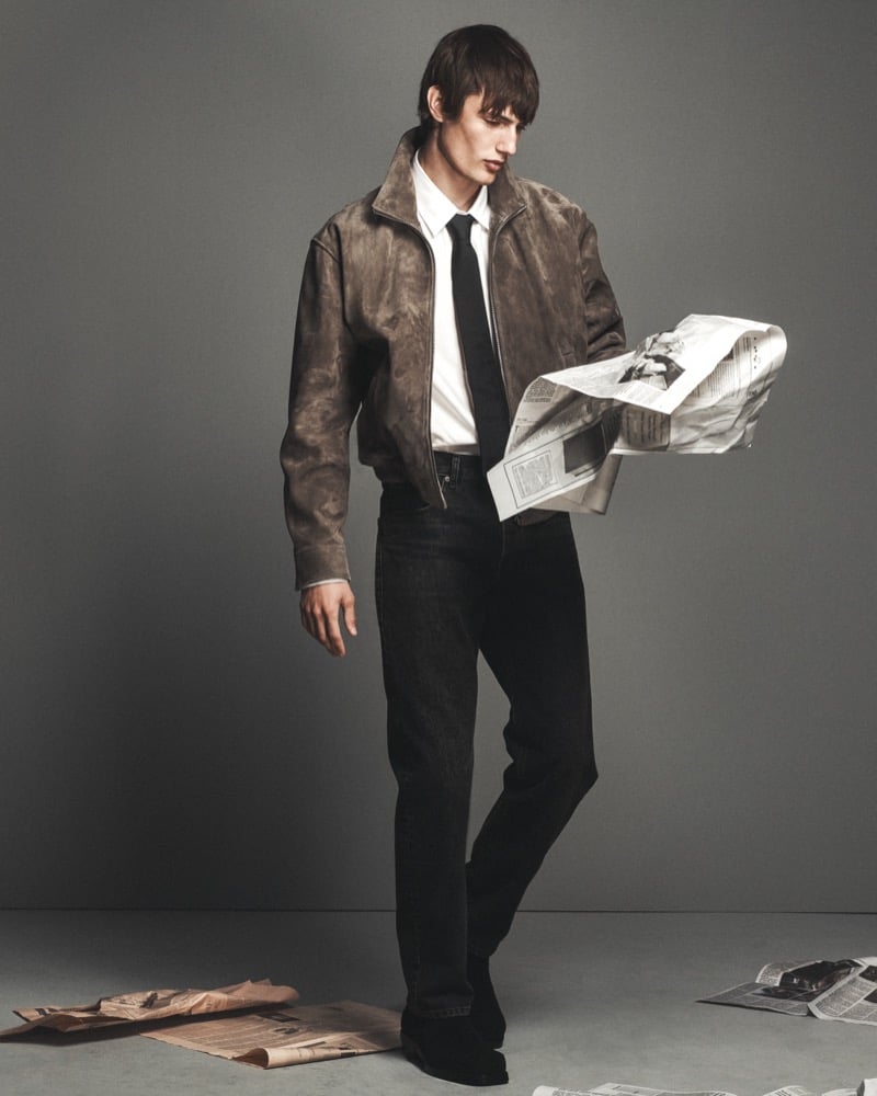 Thatcher Thornton wears a taupe suede aviator jacket over a white shirt and black tie with dark denim jeans, reading a newspaper amid scattered pages on a gray studio floor for FRAME's fall 2026 campaign