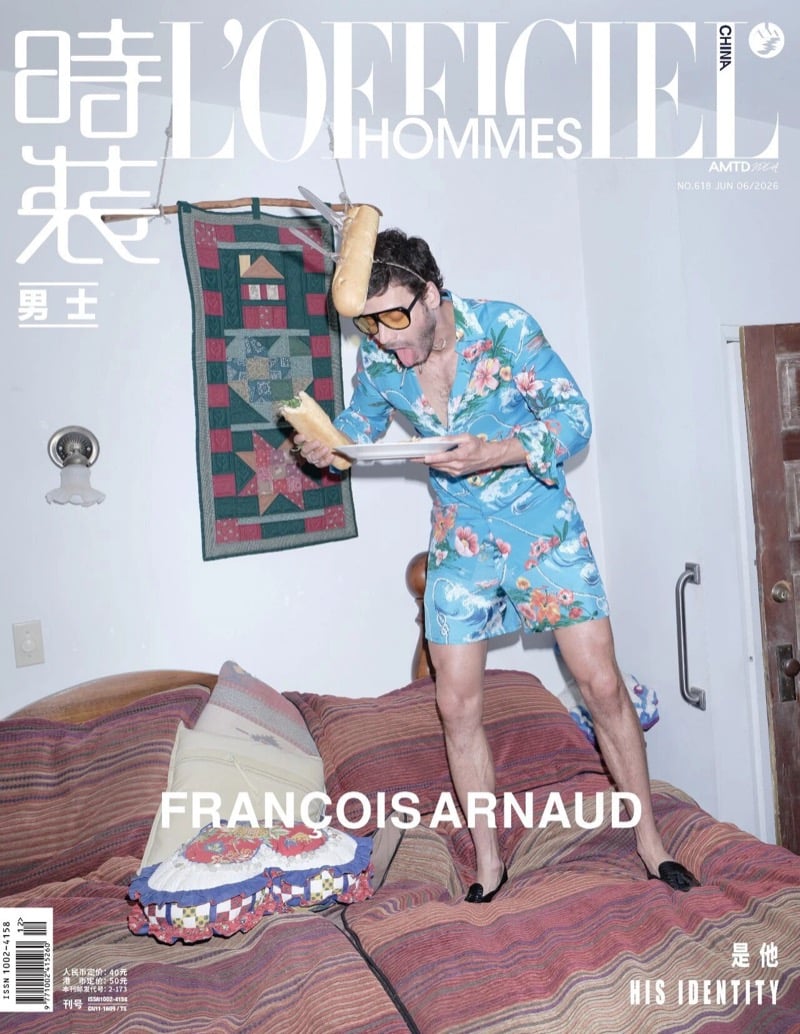 François Arnaud covers L'Officiel Hommes China standing on a bed in a blue floral pajama set, tongue out beneath a dangling baguette