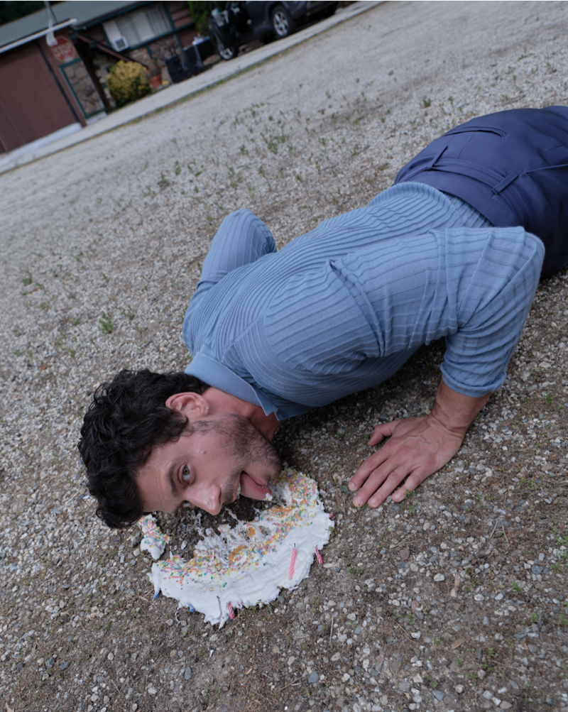 François Arnaud lies face-down in a smashed birthday cake on a gravel driveway in a blue ribbed knit shirt
