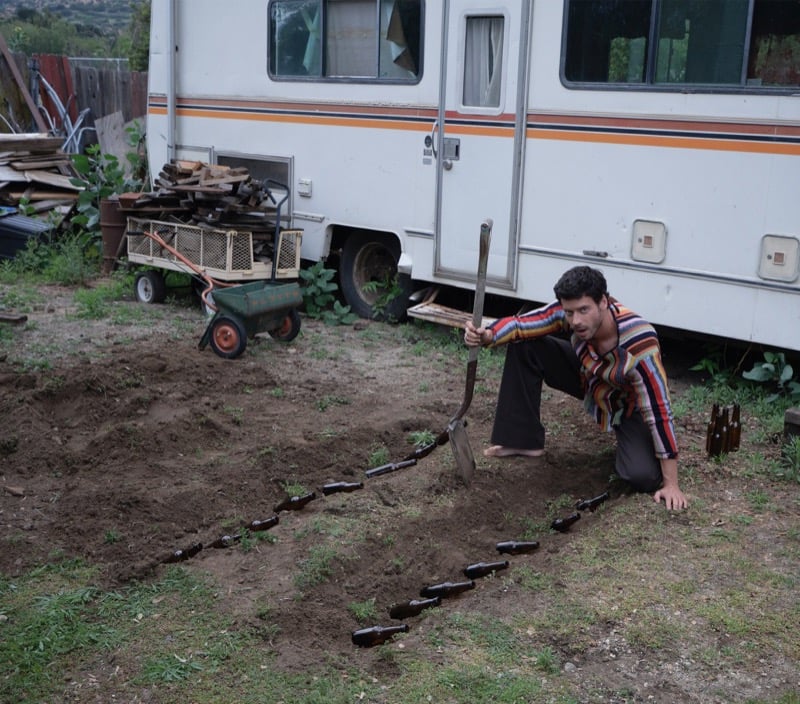 François Arnaud kneels by a vintage RV in a striped shirt, planting a row of glass bottles in the dirt with a shovel