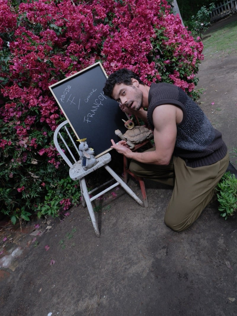 François Arnaud kneels beside a chalkboard reading F comme François surrounded by pink bougainvillea