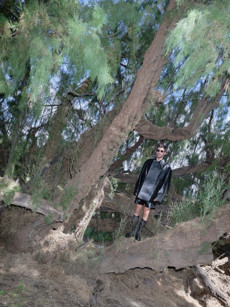 François Arnaud stands on a fallen tree branch in a black leather coat, shorts, and knee-high boots