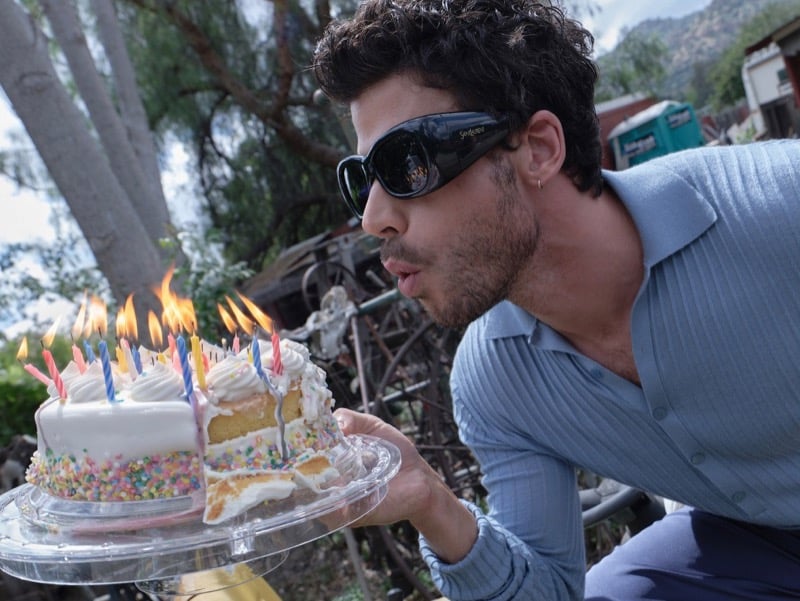 François Arnaud blows out flaming candles on a birthday cake in a close-up shot wearing dark sunglasses