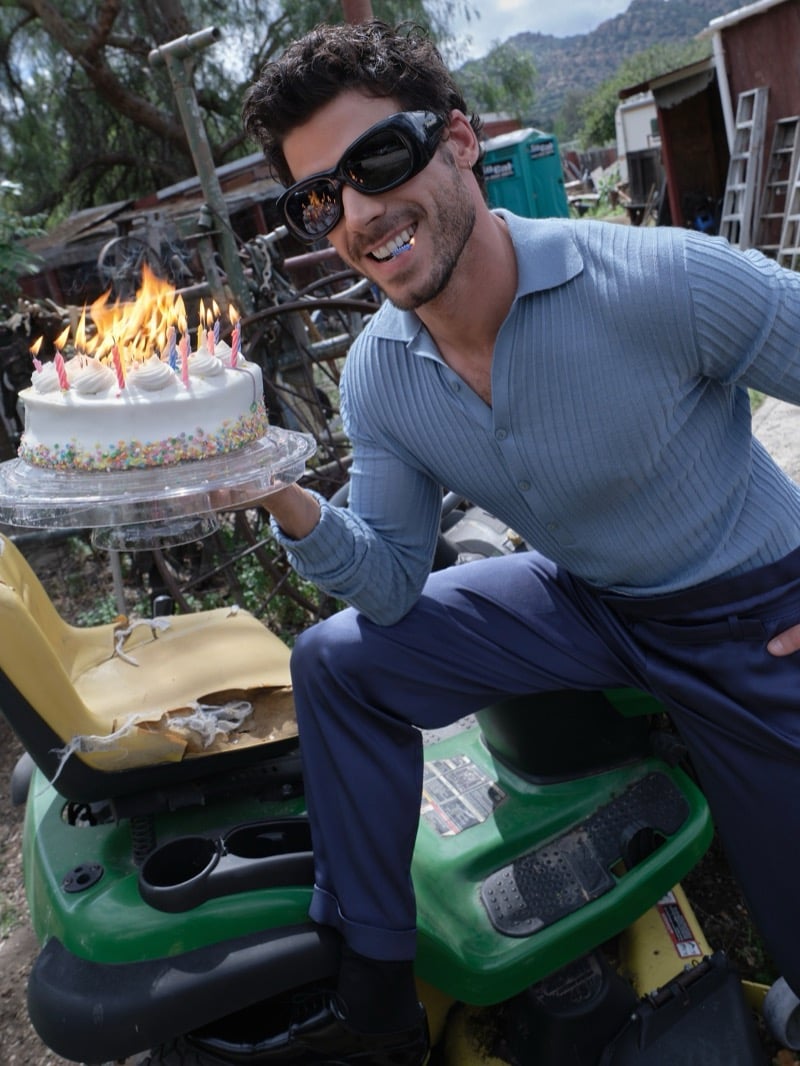François Arnaud grins on a riding lawn mower holding a lit birthday cake in a blue ribbed shirt