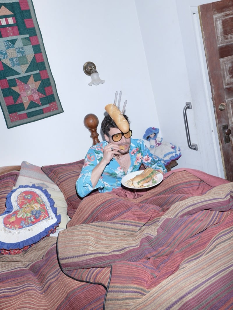 François Arnaud lies in bed in a blue floral pajama shirt with a baguette speared by forks above his head