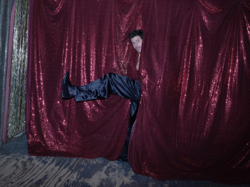 François Arnaud peeks through a maroon sequin curtain with one leg kicked out in satin trousers