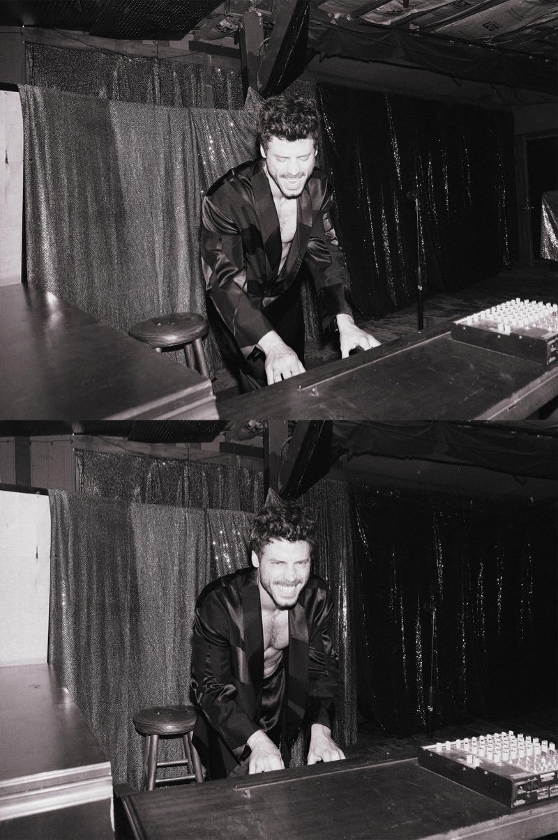 François Arnaud laughs while leaning over a piano in a black-and-white shot wearing an open satin robe