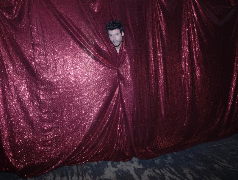 François Arnaud peers through a gap in a maroon sequin curtain, only his head visible