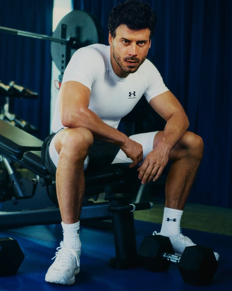 François Arnaud sits on a gym bench in Under Armour's white HeatGear compression top and shorts, dumbbells resting on the floor beside him