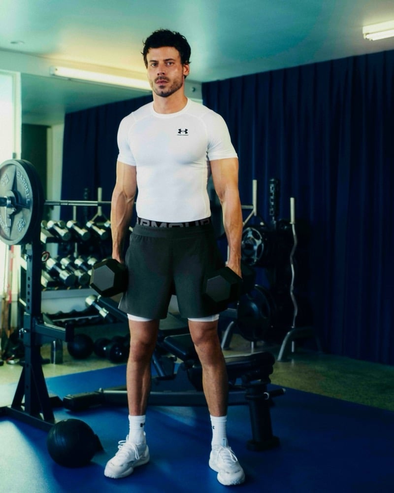 François Arnaud stands in a home gym holding a dumbbell in each hand, wearing Under Armour's white HeatGear compression top and shorts