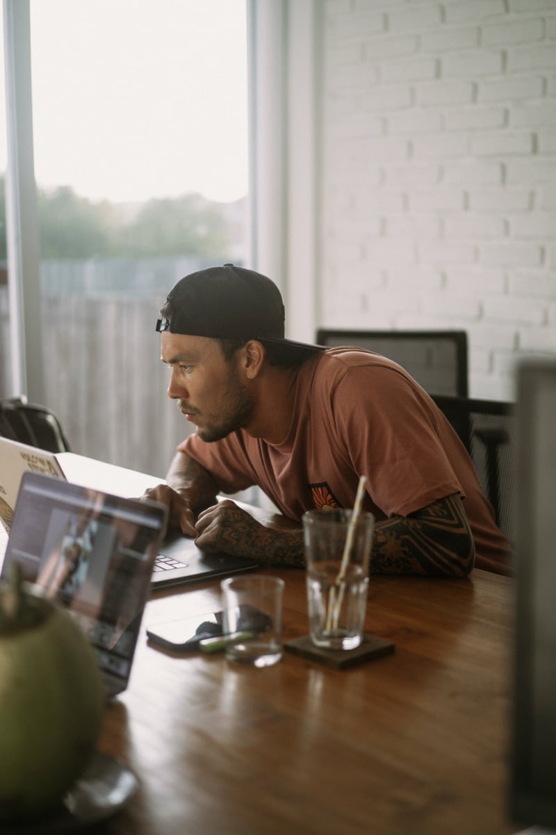 A tattooed designer works from a laptop and drawing pad at a home dining table