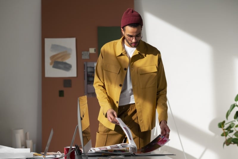 A freelance designer in a mustard jacket sorts through papers and a swatch book at a sunlit desk with dual monitors
