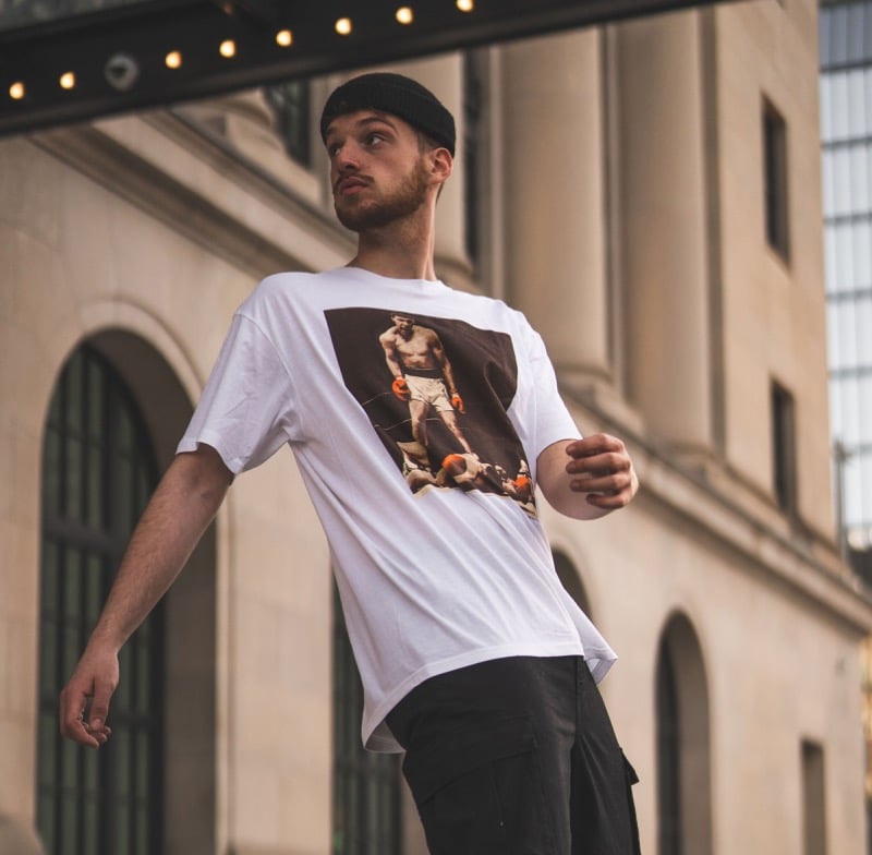 A man in a black beanie wears a white t-shirt with a photographic print on the chest