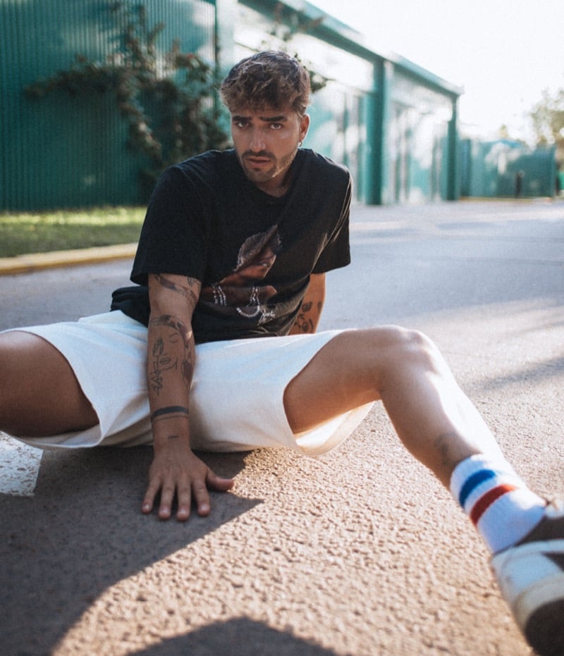 A man in a black graphic t-shirt and white shorts sits on a city street with tattoos visible on his arms