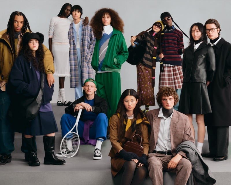 A large group of GU models in an eclectic mix of coats, knitwear, and tailoring, one seated with a tennis racket