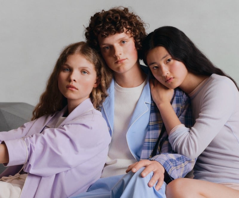 Three GU models seated closely together in a lilac jacket, a light blue jacket over a cream top, and a lavender top layered with plaid