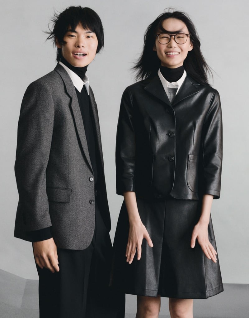 Two GU models laughing together, one in a gray blazer and dark trousers, the other in a black leather jacket and skirt with glasses