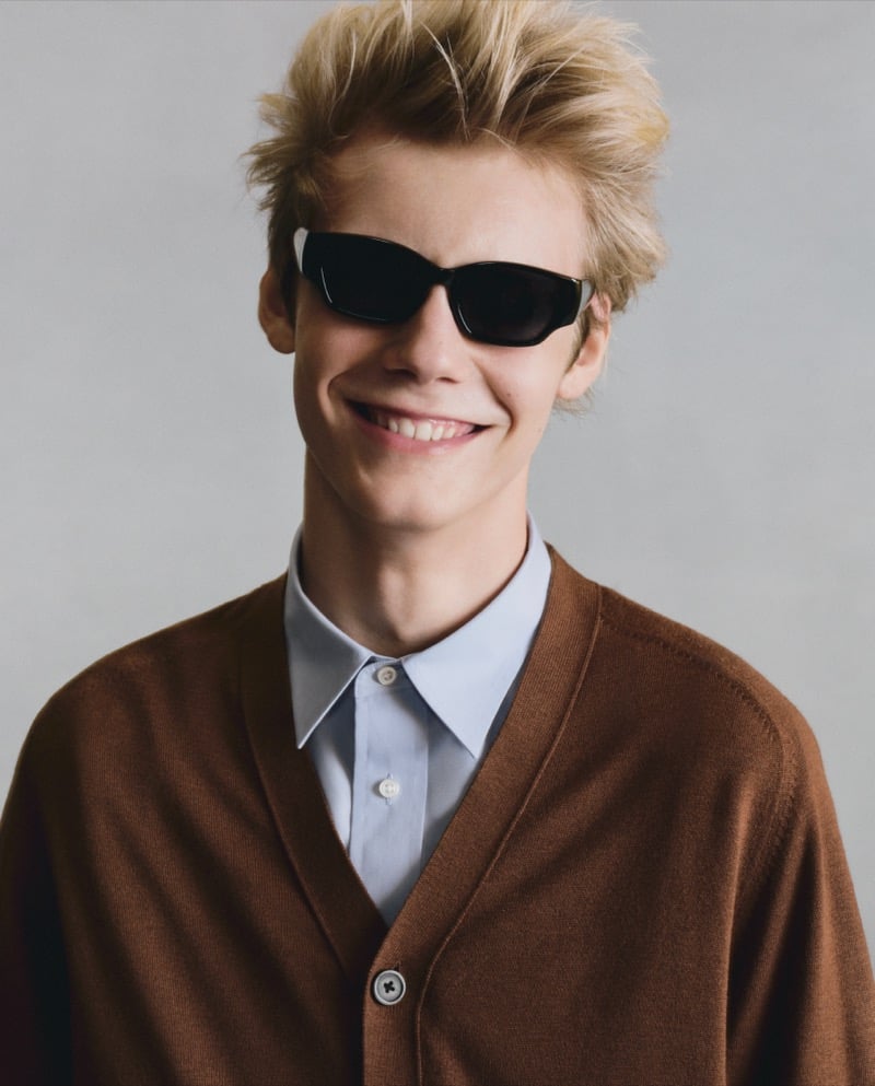 Close-up of a smiling GU model in a brown cardigan over a light blue collared shirt, wearing dark sunglasses