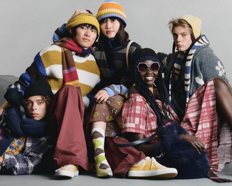 Five GU models seated together in colorful cable-knit sweaters, plaid scarves, striped beanies, and argyle socks
