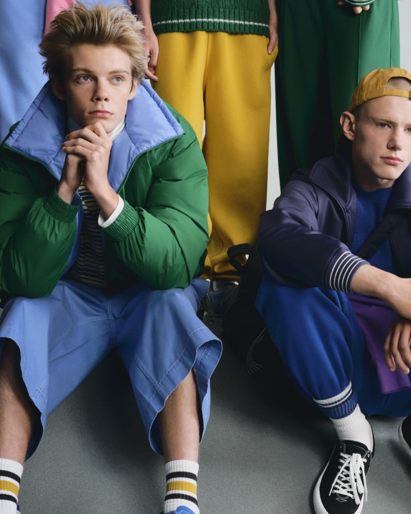 Two GU models seated, one in a green and blue puffer jacket, the other in a deep blue track jacket and yellow backward cap