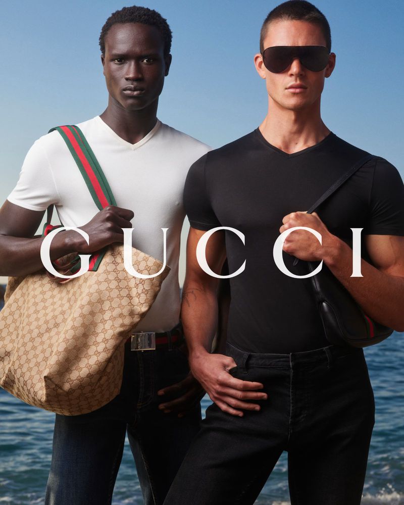Ibrahima Kane in a white v-neck holds a Gucci GG canvas tote with a green-and-red Web strap beside Samuel Watson in a black v-neck on a beach