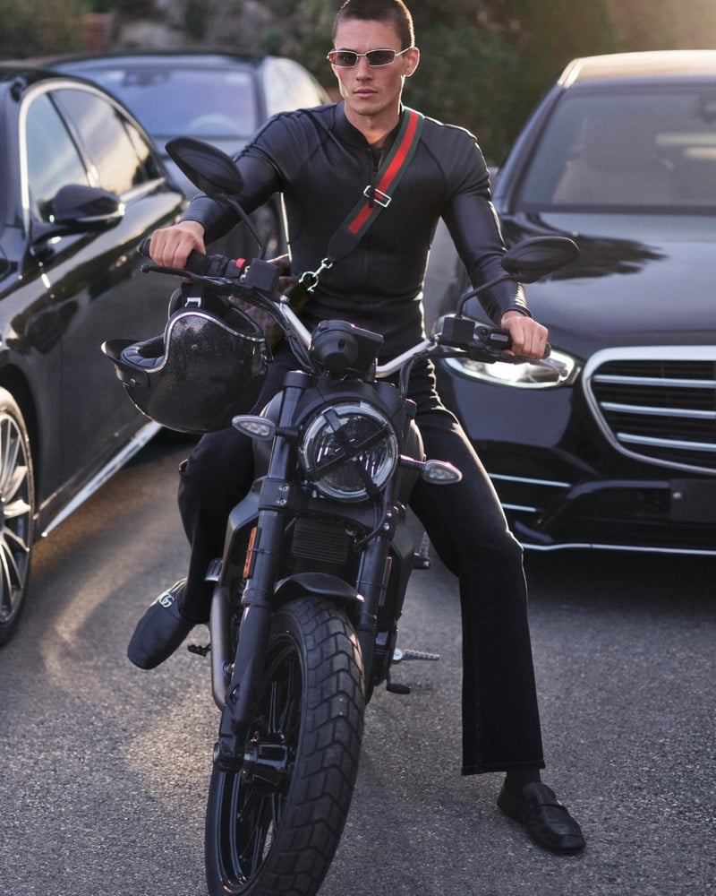 Samuel Watson rides a motorcycle in a black leather jacket with a Gucci Web-striped crossbody bag through city traffic