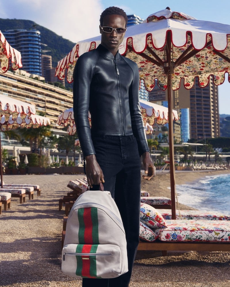 Ibrahima Kane in a black leather jacket holds a Gucci canvas backpack with a Web stripe on a beach lined with floral umbrellas