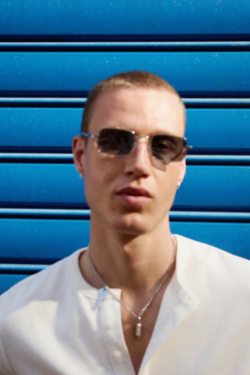 Close-up of Michael Oder wearing sunglasses and a silver necklace in an open-collar white shirt against a blue corrugated wall