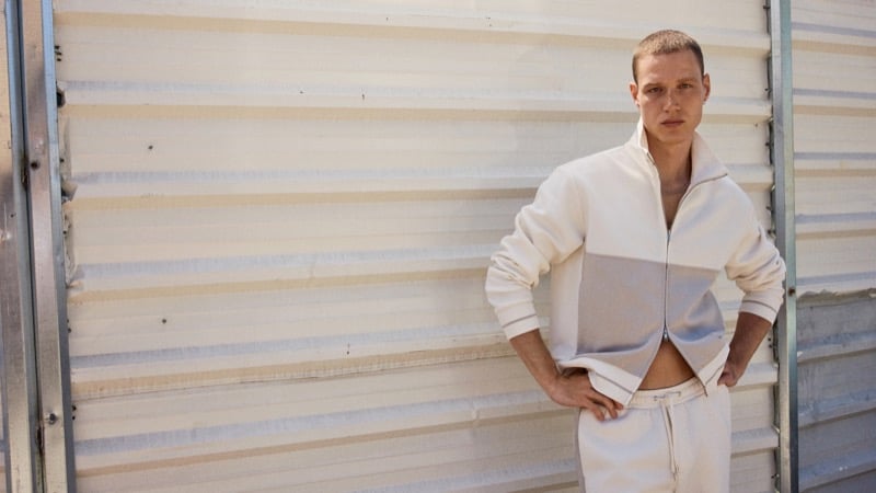 Michael Oder stands with hands on his hips wearing an ecru half-zip jacket and matching trousers against a corrugated tan wall