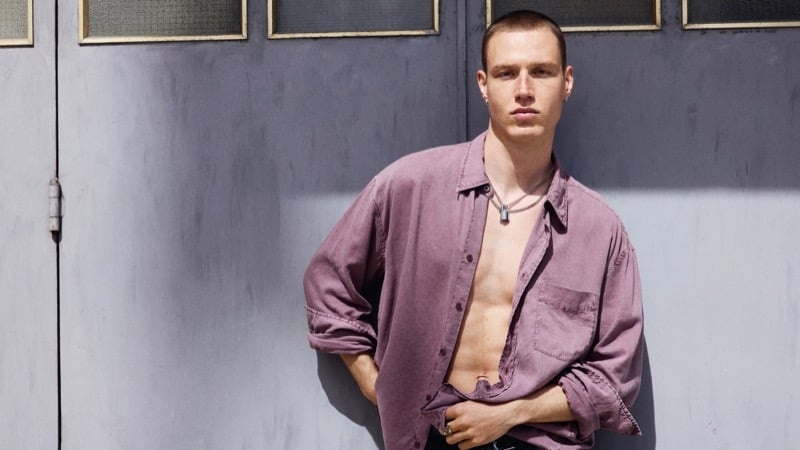 Michael Oder wears an open dusty mauve shirt with layered necklaces against a lavender-gray wall lined with framed windows