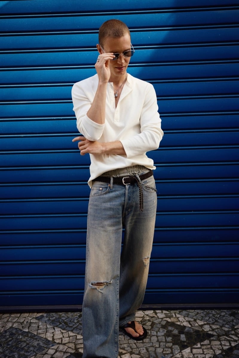 Michael Oder adjusts his sunglasses in a white popover shirt with sleeves rolled and wide faded denim, standing on cobblestones against a blue wall
