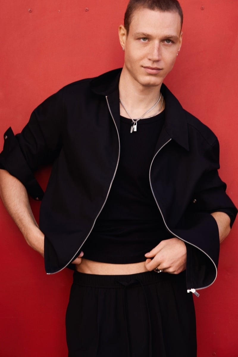 Michael Oder wears a black zip-up jacket over a black tank top and wide trousers, standing against a bold red wall