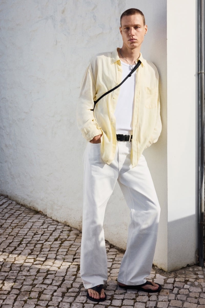 Michael Oder wears an open butter yellow shirt over a white tank top with wide white jeans, a black crossbody strap, and flip-flops on a cobblestone street