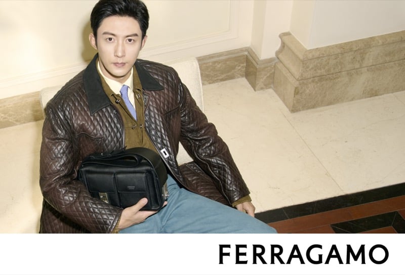 Huang Jingyu sits wearing a quilted brown leather jacket with a dark collar, holding a black structured bag in his lap